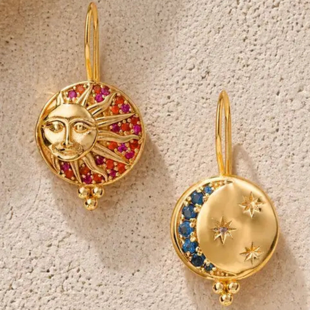 A pair of gold celestial drop earrings displayed on a textured beige surface. One earring features a gold sun face surrounded by red and pink gemstones, and the other features a gold crescent moon and stars set with blue and clear gemstones.